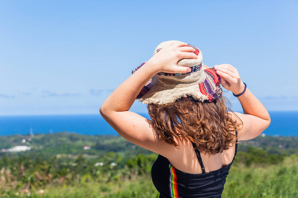 Νεαρό κορίτσι γυναίκα στις διακοπές closeup πίσω από την πλάτη τους ώμους τα χέρια, κρατώντας το καπέλο από τους ανέμους που αναζητούν πάνω από το μακρινό μπλε του ωκεανού και πράσινο καλοκαίρι ύπαιθρο ακτογραμμή. - Φωτογραφία, εικόνα