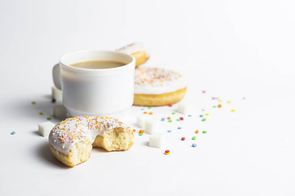 Bílý hrnek, káva nebo čaj s mlékem a čerstvé chutné koblihy, sladké barevné Dekorativní cukroví na bílém pozadí. Koncept pekárny, čerstvé pečivo, vynikající snídaně, rychlé občerstvení - Fotografie, Obrázek