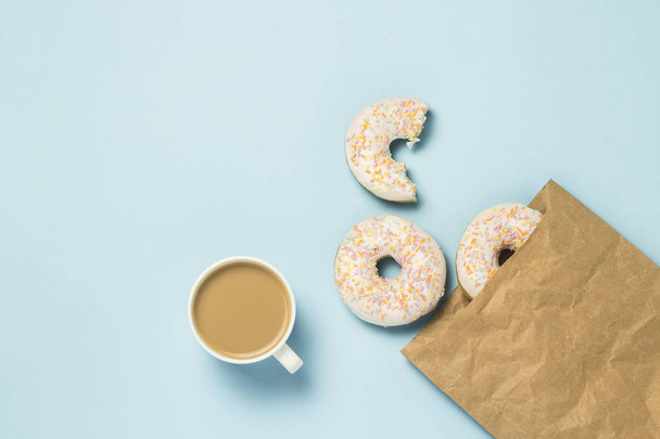 weiße Tasse mit Kaffee oder Tee und Papiertüte mit frischen leckeren süßen Donuts auf blauem Hintergrund. das Konzept von Fast Food, Bäckerei, Frühstück, Süßigkeiten. Minimalismus. flache Lage, Draufsicht - Foto, Bild