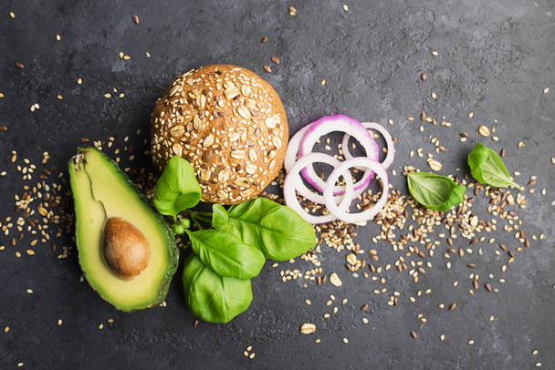 Ingredients of burger with avocado. Vegetable burger made from wholegrain bun, avocado, sweet onions, cherry tomatoes, basil on a dark background. Top view, space. - Photo, image