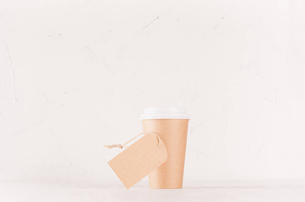 Coffee packing mockup -  craft brown paper cup with white cap and blank label on white wood board, coffee shop interior. Modern elegant concept for branding identity, advertising, design. - Φωτογραφία, εικόνα