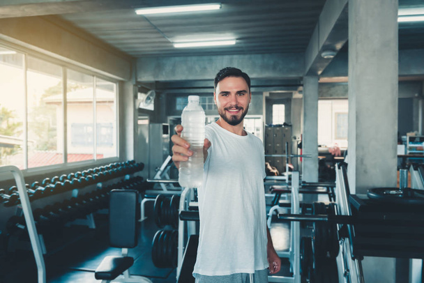 Portrait of sporty male holding bottle of water to giving someone in front of camera on exercise equipment background., Smiling sports man working out in gym.,Sport club and healthy concept. - Photo, Image