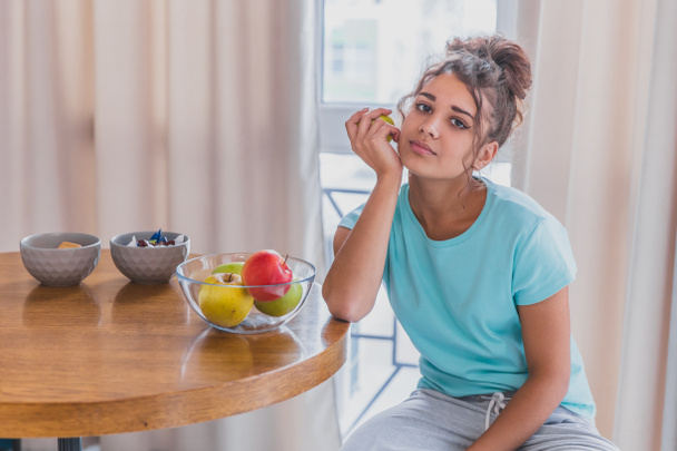 Ευτυχισμένη γυναίκα δροσιστικό με φλιτζάνι καφέ, σνακ και φρέσκα φρούτα στο τραπέζι της κουζίνας, νωρίς το πρωί. - Φωτογραφία, εικόνα