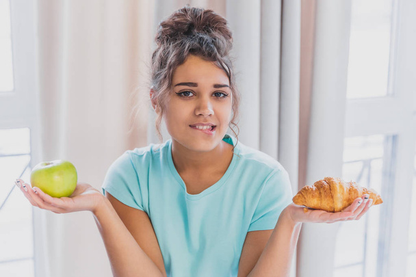 Beautiful young slender brunette with green apple in one hand and croissant in the other hand on background of vegetables on kitchen table making a difficult choice between healthy and harmful food. - Foto, immagini