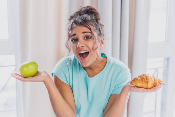 Beautiful young slender brunette with green apple in one hand and croissant in the other hand on background of vegetables on kitchen table making a difficult choice between healthy and harmful food. - Fotoğraf, Görsel