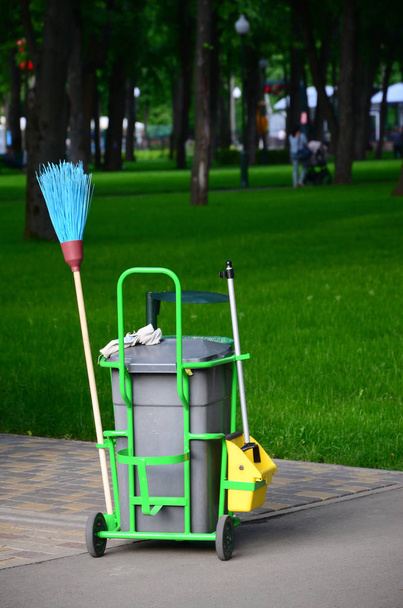 Carro de servicio de limpieza lleno de suministros y equipos junto con contenedor de basura gris para limpiador profesional contra hierba verde en el parque público
 - Foto, Imagen