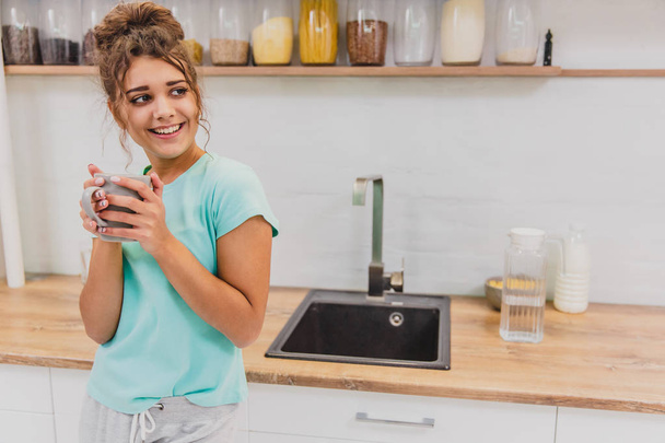 Gorgeous, beautiful, young woman holding glass of fresh water and smiling while standing in kitchen in the morning - Foto, imagen