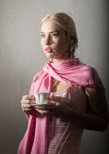 junge blonde Frau hinter Glas mit Wassertropfen. schönes Mädchen trinkt Kaffee oder Tee - Foto, Bild
