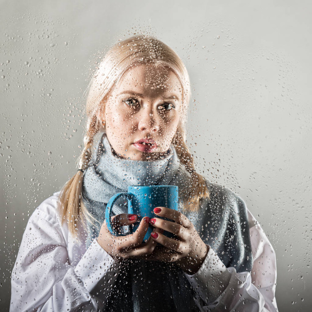 young blonde woman behind glass with water drops. beautiful girl drinks coffee or tea - Φωτογραφία, εικόνα