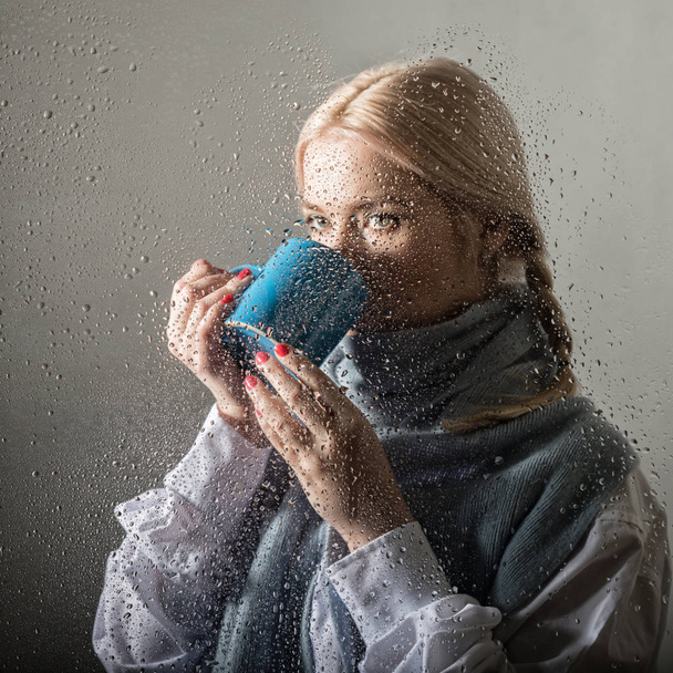 young blonde woman behind glass with water drops. beautiful girl drinks coffee or tea - 写真・画像