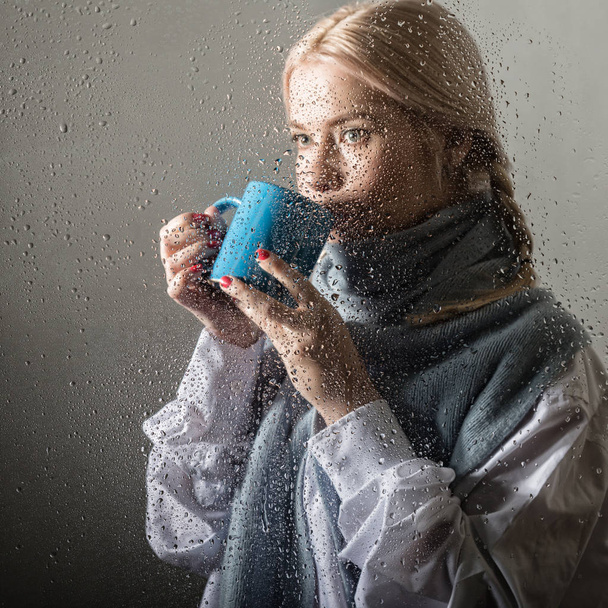 young blonde woman behind glass with water drops. beautiful girl drinks coffee or tea - Foto, Bild