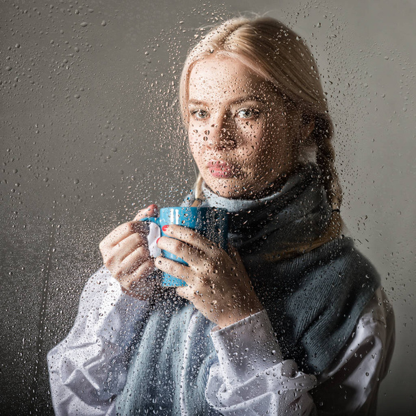 young blonde woman behind glass with water drops. beautiful girl drinks coffee or tea - Φωτογραφία, εικόνα