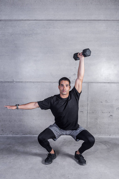 Young sportive trainer holding dumbbell and doing shoulder press in squat position on gray concrete  - Photo, Image