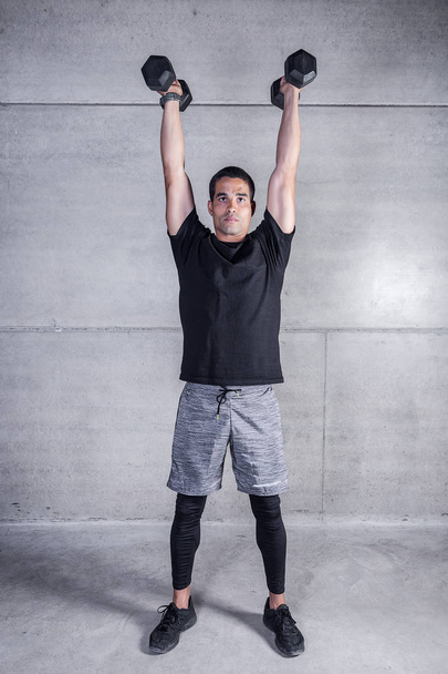 Focused sportive man holding arms with dumbbells raised above head standing on gray concrete  - Fotografie, Obrázek