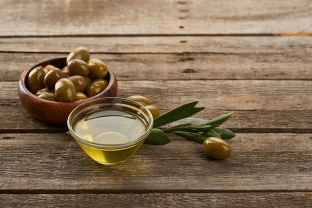 glass bowl of oil, wooden bowl with olives, olive tree leaves and olives on brown wooden surface - Foto, Bild