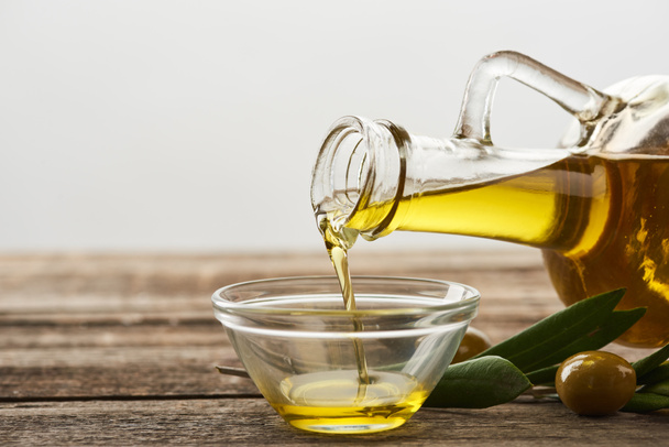 vylévání oleje z láhve do skleněné mísy, listy olivovníku a olivy na dřevěný povrch - Fotografie, Obrázek