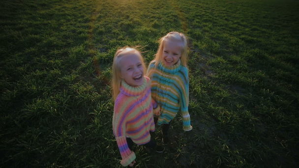 slow motion happy twin sisters in loose striped clothes run to camera and laugh on green meadow in evening - Footage, Video