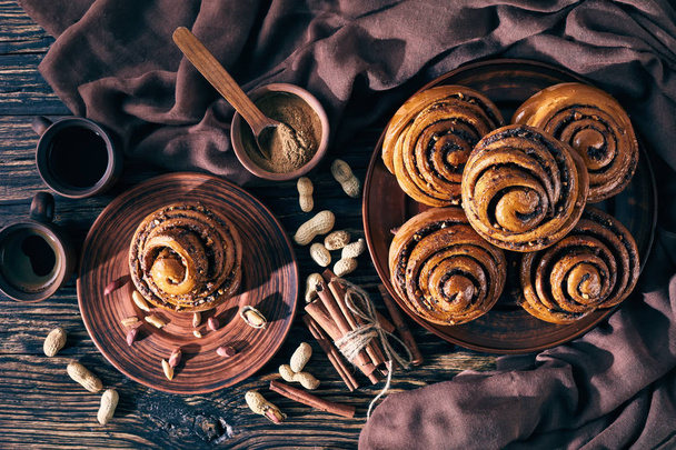 Cinnamon rolls buns with peanuts on a earthenware plate with brown cloth and coffee on a rustic table. Kanelbulle swedish dessert, view from above, flatlay - Photo, Image