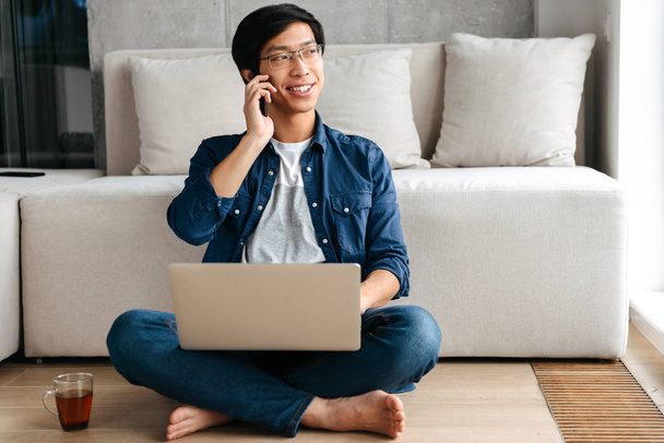 Szczęśliwy człowiek asian siedzi na kanapie w domu, przy użyciu komputera przenośnego, rozmowy na telefon komórkowy - Zdjęcie, obraz