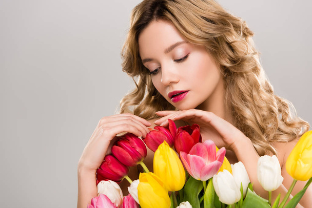 グレーに分離されたカラフルなチューリップの花束に触れる魅力的なばねの若い女性  - 写真・画像