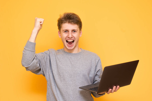 Emotional young man in the casual clothing holds a laptop in his hands, looks in camera and rejoices with a raised hand on a yellow background. Portrait of a joyful student man on a colored background - Photo, Image
