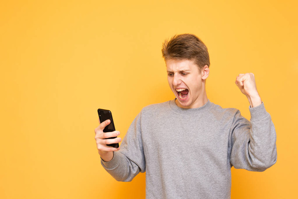 Expressive guy in casual clothing looks at the smartphone screen and rejoices, hands up on a yellow background. Joyful young man with a smartphone in his hands is isolated on a yellow background. - Foto, afbeelding