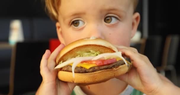 Szép boldog éhes baby boy eszik hamburgert. Fast food koncepció. Ízletes egészségtelen hamburger szendvics kezében - Felvétel, videó