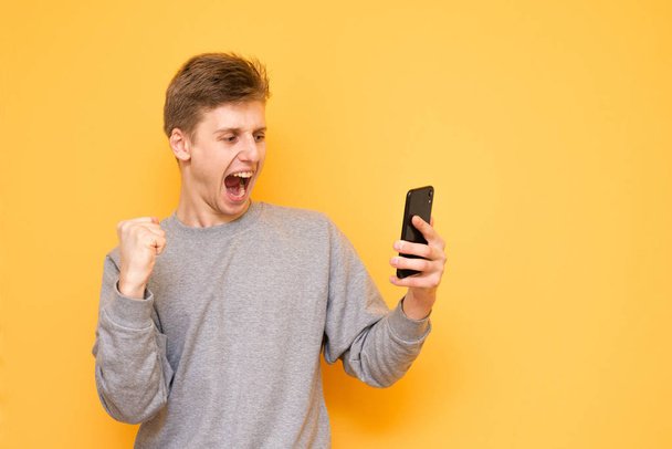 Gamer gamer plays a game on a smartphone,looks at the smartphone screen and makes a gesture to the winner.Emotional gamer with a smartphone in his hands plays game on a yellow background and rejoices - Foto, afbeelding