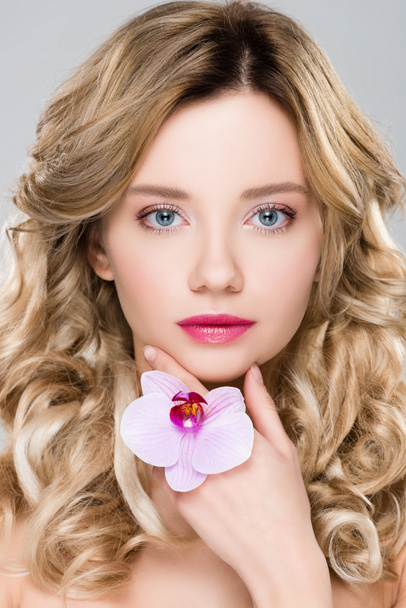 young beautiful woman holding purple orchid and looking at camera isolated on grey - Photo, Image