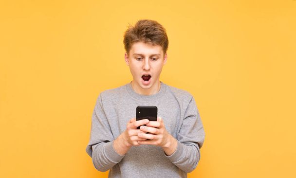 Portrait of an emotional guy standing on a yellow background, using a smartphone and looking surprised at the screen.Amazed young man uses the Internet on a smartphone, isolated on a yellow background - Fotografie, Obrázek