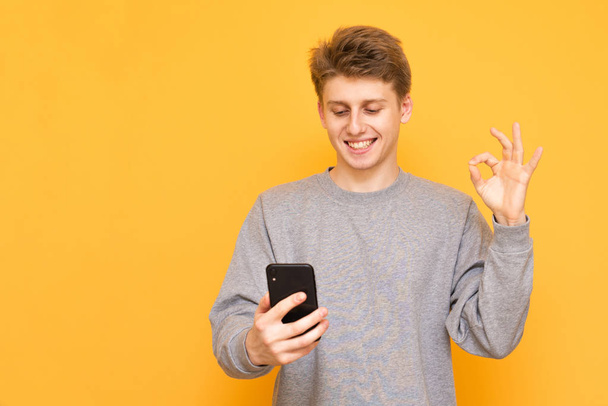 Positive guy uses a smartphone on a yellow background, looks at the screen and shows the OK sign. Happy young man with a smartphone in his hand is isolated on a yellow background - Foto, imagen