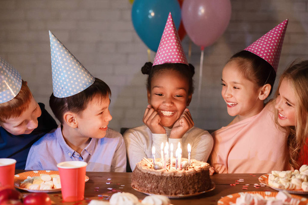 Діти святкують день народження вдома, дивлячись на торт зі свічками
 - Фото, зображення