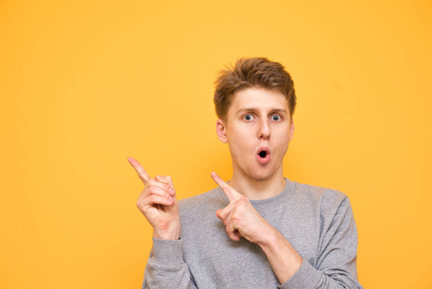 Smiling young guy is standing on a yellow background and showing his fingers on copyspace. Positive man shows his hands aside and looks at the camera, isolated on a yellow background. - Фото, изображение
