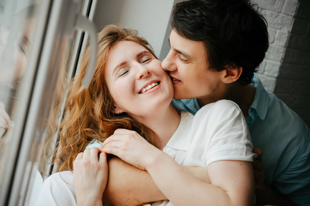 Ευτυχισμένος κορίτσι και man φιλιά κοντά στο παράθυρο στο σπίτι.  - Φωτογραφία, εικόνα