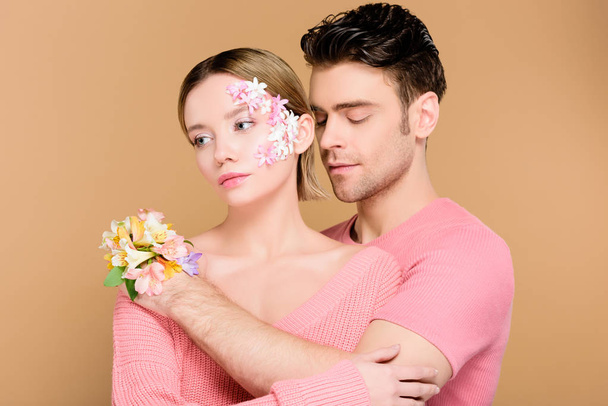 pohledný muž všeobjímající atraktivní přítelkyni s květinami na tváři izolované na béžové  - Fotografie, Obrázek