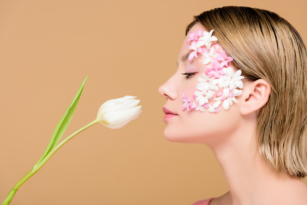 side view of elegant woman with flowers on face smelling tulip isolated on beige - Foto, Imagem