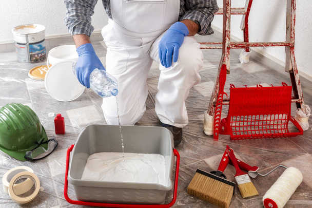Pintor de casa caucásico trabajador en mono de trabajo blanco, vierte el agua en el recipiente con la pintura. Industria de construcción
. - Foto, imagen