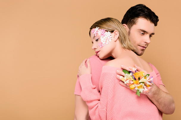 handsome man with alstroemeria flowers on hand embracing girlfriend with flowers on face isolated on beige  - Foto, Imagen