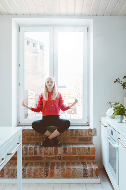 Blond young woman wearing sport clothes doing yoga by the window at home.  - Valokuva, kuva