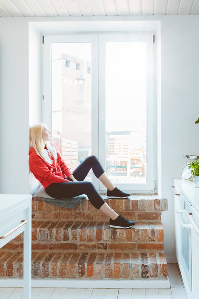 Blond young woman wearing sport clothes resting by the window at home.  - Foto, Bild