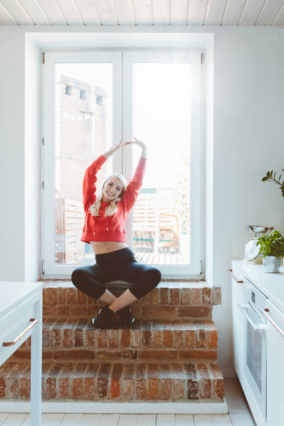 Blond young woman wearing sport clothes stretching by the window at home.  - Фото, изображение