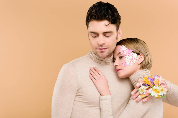 tender woman with flowers on face hugging handsome man isolated on beige  - Foto, Bild