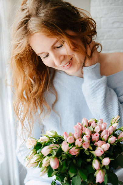 Portrait of a young beautiful girl with a bouquet of roses near window. Curly flowing red hair. - Zdjęcie, obraz