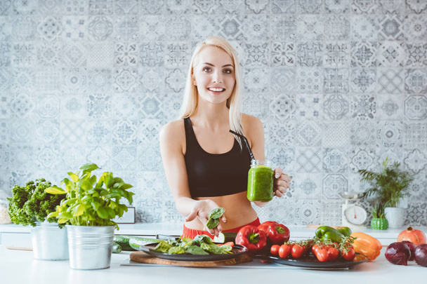 Blonde young woman wearing sport clothes preparing green smoothie in the kitchen, holding drink and spinach in hands, smiling at camera.  - Photo, image