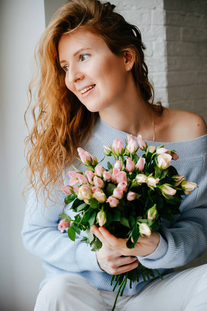 Portrait of a young beautiful girl with a bouquet of roses near window. Curly flowing red hair. - Photo, Image