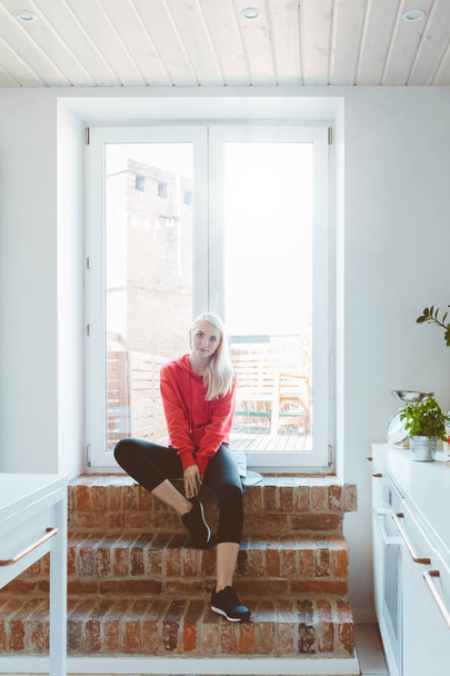 Blond young woman wearing sport clothes resting by the window at home.  - Foto, Imagen