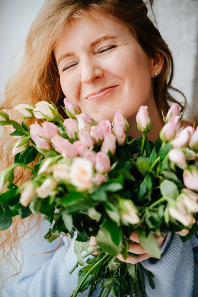 Portrait of a young beautiful girl with a bouquet of roses near window. Curly flowing red hair. - Foto, Bild