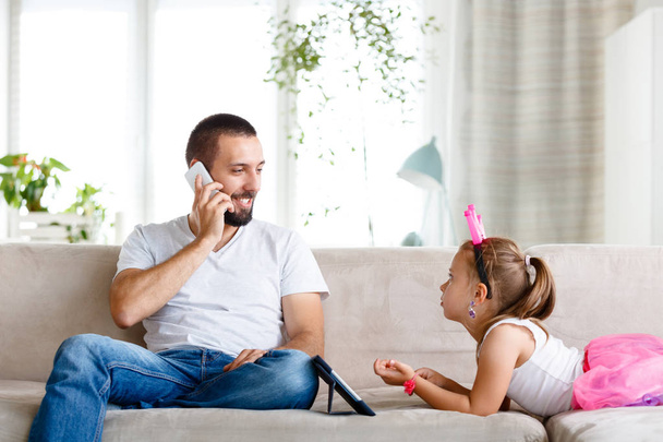 Νεαρός κάθεται στον καναπέ στο σαλόνι και να μιλάτε στο τηλέφωνο, ενώ η κόρη του βλέποντας ψηφιακή δισκίο.  - Φωτογραφία, εικόνα
