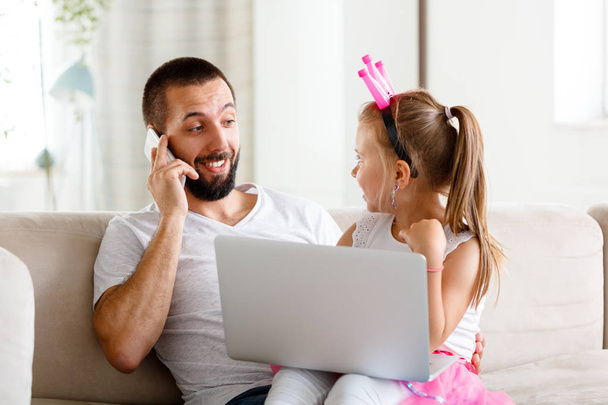 Father and daughter spending time together at home. Young man talking on phone while his cute daughter using laptop.  - Φωτογραφία, εικόνα