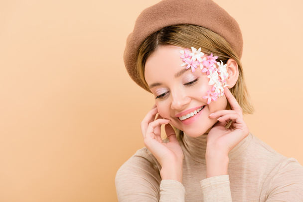 cheerful with flowers on face wearing trendy beret isolated on beige  - Zdjęcie, obraz